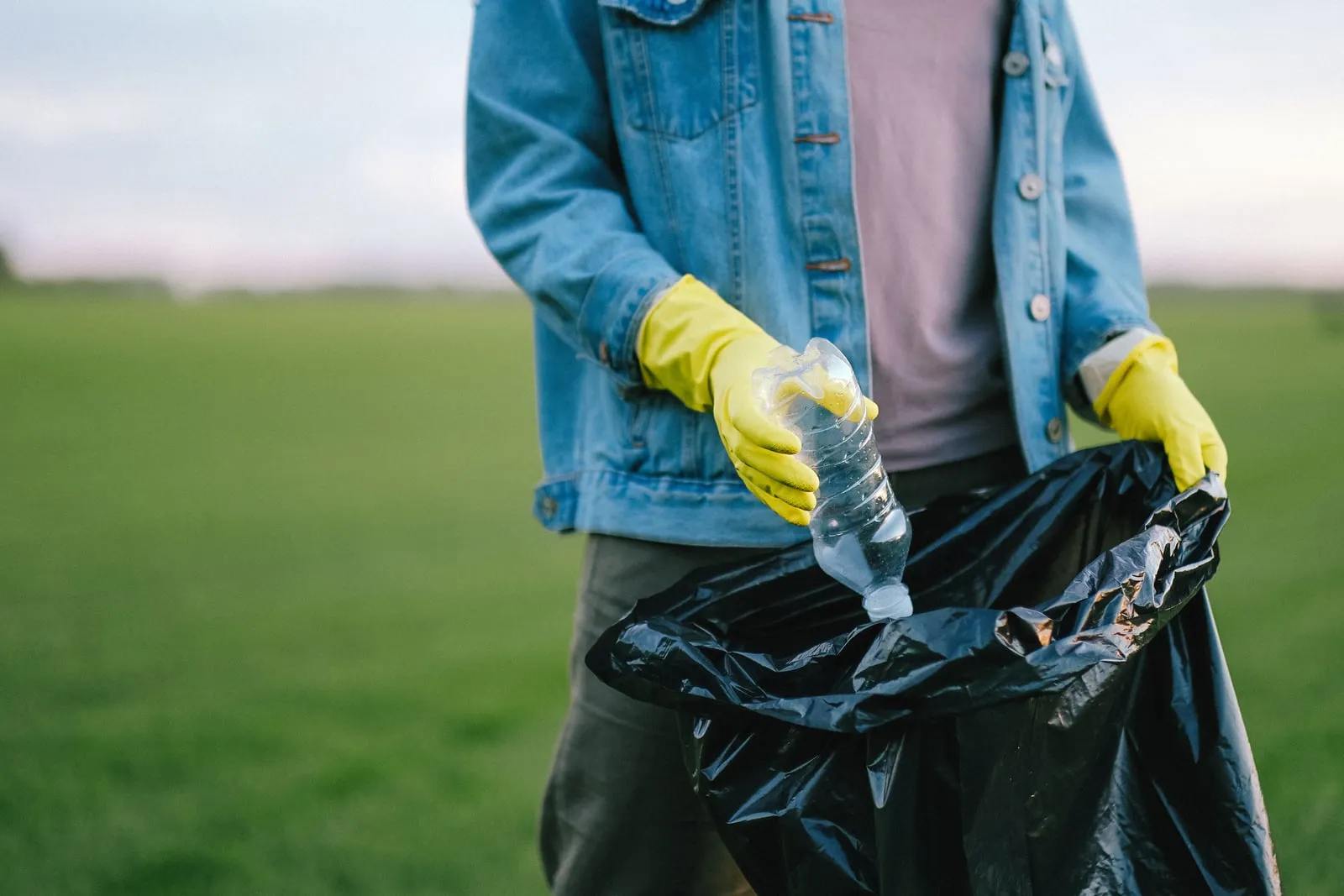 A gloved hand drops a plastic bottle into a black trash bag in an open green field, cleaning up garbage at the source